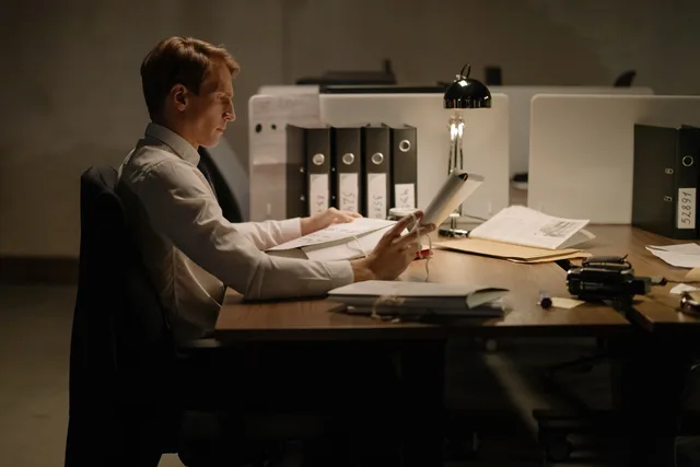 An office worker in formal attire intensely focused while reading documents at a desk with folders and a lamp. PR: så bygger du förtroende genom redaktionell synlighet